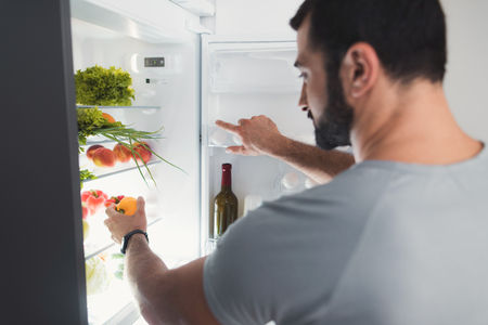 A sporty man stands in the kitchen and takes fresh vegetables from the refrigerator.の写真素材