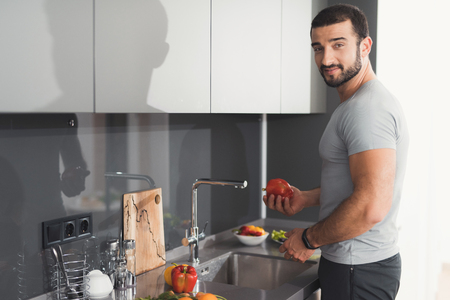 A sporty man is standing in the kitchen and washes vegetables for a salad.の写真素材