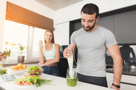 Young sports couple is preparing in the morning in the kitchen. A man is making a green vegetable cocktail.の写真素材