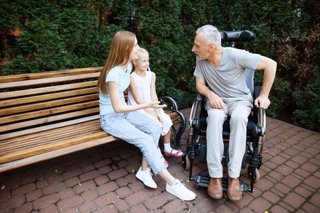 An elderly man is sitting in a wheelchair. He is seen by a woman with a girl. They are sitting on a bench in the park.の写真素材