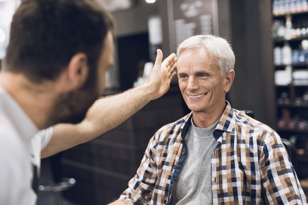 The old man is sitting in the barbers chair in a mans barbershop, where he came to cut his hair.の写真素材