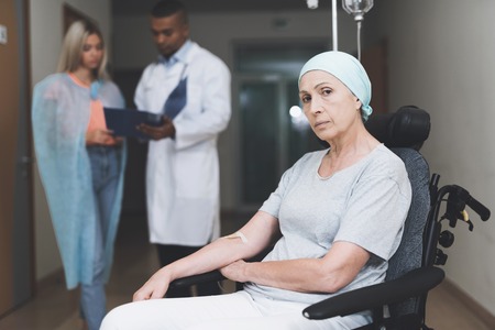 A woman with cancer, with a doomed look, is sitting in a wheelchair. Behind her daughter stands and...