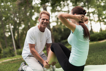 A woman is rocking the press on nature. She sits on a rug for yoga. A man helps her and holds her legs.の写真素材