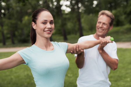 Girl doing exercises with dumbbells in the park. A man helps her. They are smilingの写真素材