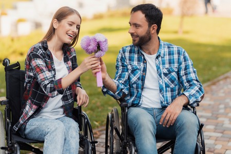 A couple of invalids on wheelchairs met in the park. A man is giving a bouquet of flowers to a woman.の写真素材