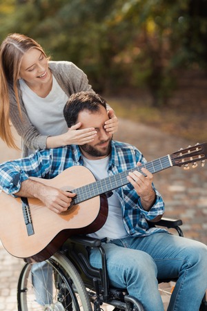 A man on a wheelchair playing guitar in the park. A woman came to him from behind and closed her eyes with her hands.の写真素材