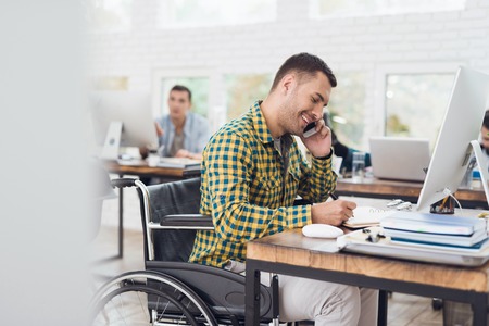 A man in a wheelchair writes with a pen in a notebook and talks on the phone. He is working in a bright office.の写真素材