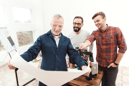 Three engineers work with a 3d printer. An elderly man in the foreground is studying a blueprint.