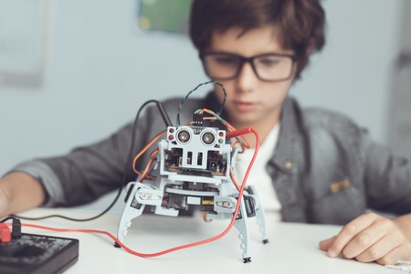 A small nerd in glasses is holding a robot. He looks carefully at his creation