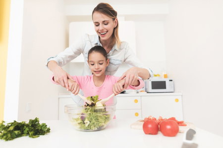 Mom and daughter have fun while preparing a salad. They are in a bright kitchen.の写真素材