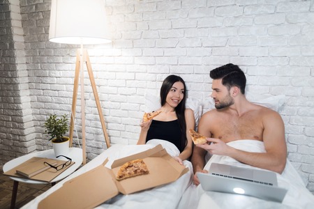 Guy is lying in bed and working on laptop. Girl is sitting near and they are eating pizza in bed.の写真素材