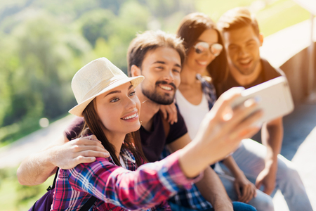 A group of tourists are sitting in a park on a bench. They do selfie on a smartphone.の写真素材