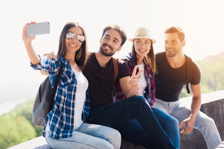 A group of tourists are sitting in a park on a bench. A girl in glasses makes selfie with her...