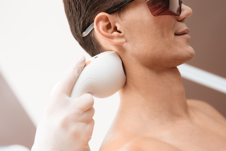 The man came to procedure of laser hair removal. The doctor treats his shoulders, neck and back with a special apparatusの写真素材