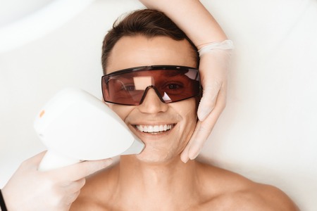 The doctor treats the face of a man with a modern laser epilator. The man lies and smiles.の写真素材