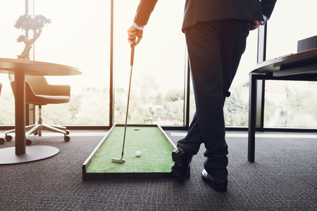 Close up. A man in a business suit playing golf in the office. He is playing on a green mat.