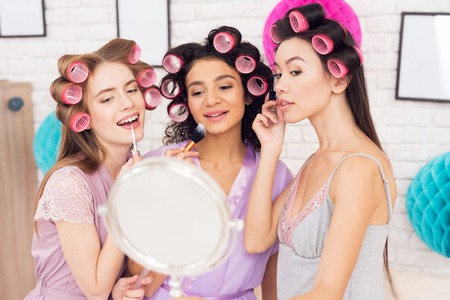 Three girls with curlers in their hair doing make up behind mirror. They are celebrating womens day March 8.の写真素材