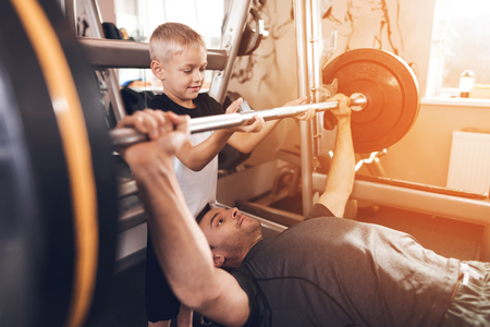 Father and son in the gym. Father and son spend time together and lead a healthy lifestyle. The father lifts the barbell and the son helps him.の写真素材
