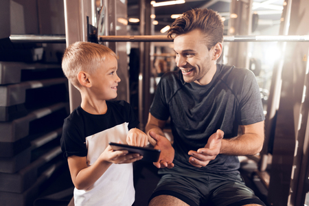 Father and son in the gym. Father and son spend time together and lead a healthy lifestyle. A boy shows something to his father on a tablet in the gym.の写真素材