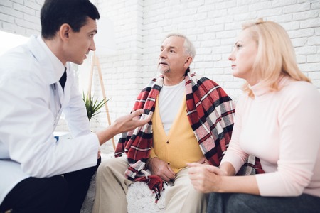 A doctor came to the old man in a yellow cardigan. The old man became ill and the doctor diagnoses him. Next to him is the old man's wife. They are in the house of an elderly couple.の写真素材