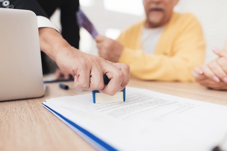 Mature couple at a reception with a lawyer. The lawyer puts a seal on the contract, the old people are happy about this. They are sitting at the desk in the lawyer's office.の写真素材