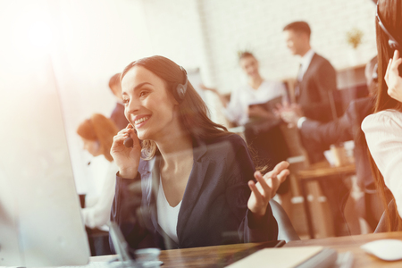 The brunette girl is sitting at the computer. She works as a call center operator. She communicates with customers by phone.の写真素材