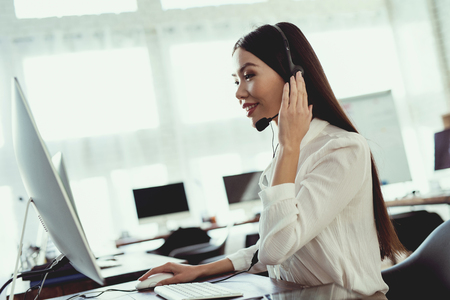 Asian girl sitting in the call center. She has headphones on which she talks to customers. There is a computer in front of her.の写真素材
