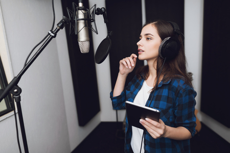 The girl in the recording studio sings a song. She has headphones on her head, and a tablet with text in her hands. Next to her is a microphone. She emotionally sings the song.の写真素材