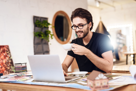 Freelancer bearded man in t-shirt drinking tea at laptop sitting at desk.の写真素材
