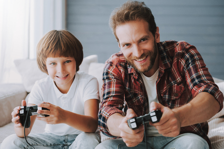 Cheerful father with game controller plays video game with small son sitting on couch. Parent spends...