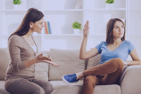 Teen girl showing stop gesture to angry mother while sitting on sofa at home.Problems in family concept.の写真素材