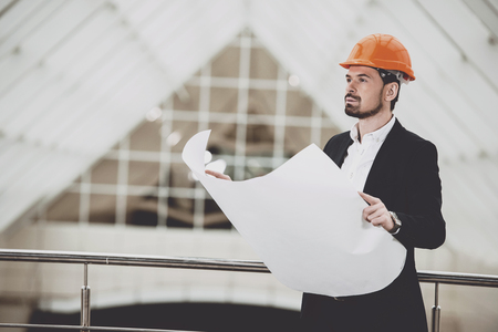 Photo of male architect in red hardhat holding blueprints and looking at building under construction.の写真素材