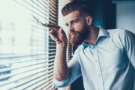 Young and Handsome Businessman in White Shirt near Windows in Modern Office. Bearded Young Man near Jalousie. Office Worker at Work. Business Concept. Work in Modern Office Concept.の写真素材