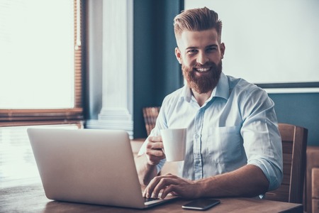 Young and Handsome Businessman in White Shirt with Laptop and Cup of Coffee in Modern Office. Happy Office Worker at Work. Communication at Work Concept. Business Concept. Guy with Gadget.の写真素材