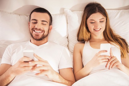 Young Smiling Couple Lies in Bed and Watching into Smartphones. In Morning after Sleep. Love and Relationships Concepts. Family Life Concept. Idyll of Young Family. Using Gadgets in Bed.の写真素材