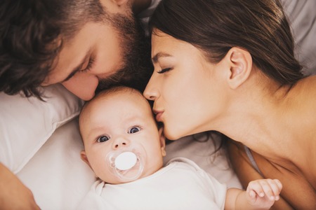 Top view. Mother and Father Kisses Baby in Bed. Little Newborn Child in White Bed. Parenthood Concepts. Love and Happiness. Taking Care of New Life. Family, Love, Happiness Concepts.の写真素材