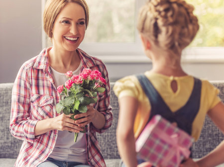 Mother Holding Flowers and Looking at Her Daughter. Beautiful Mom Smiling While Sitting on Soft Couch at Sweet Home. Little Cute Girl Holding Gift Box Behind Her Back. Family Holiday and Togetherness.の写真素材