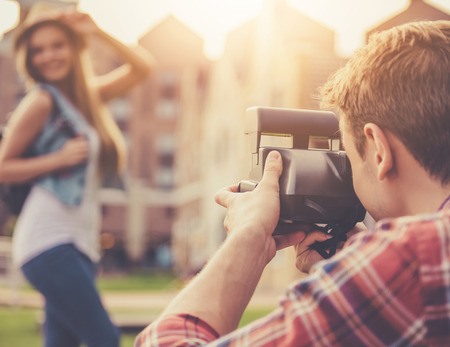Handsome Guy Takes Pictures of Attractive Girl in Casual Clothes and Hat. Beautiful Girl Posing and Smiling. Professional Handsome Photographer Enjoys Magic Moment. Sunny Evening.の写真素材