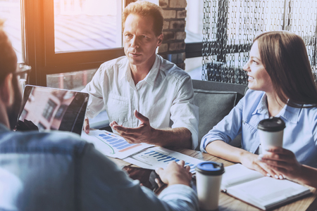 Business People in Casual Wear Discussing Affairs. Four Smart Colleagues Using Laptop and Drinking Coffee. They Have Discuss Hard Project. Professional relationships Concept. Modern Free Workplace.の写真素材