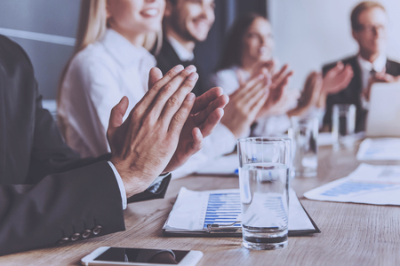 Smiling People Clap Their Hands at Conference. Mobile Phone and Glass of Water Closeup. Blur Image Inside Conference Room as Background. Businessmen in Formal Wear Participating in Conference.の写真素材