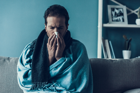 Bearded Man with Flue Sitting on Sofa at Home. Comfortable Apartment. Bearded Man in Blue Blanket. Modern Interior. Sofa witn Pillows. Illness Concept. Relaxation at Home. Healthcare Concept.の写真素材