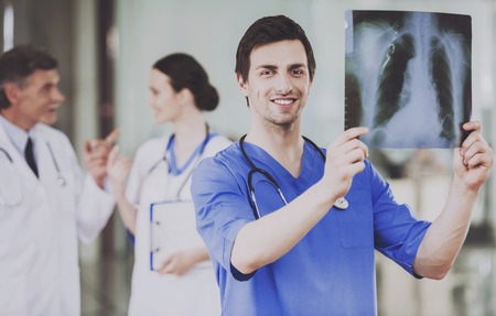 Smiling Surgeon Carefully Examine X-ray Scan in Hospital. Work in Clinic. Concept of Health Care. Medical Consultant. Holding Scan. Clinical Concept. Standing in Hospital. People in Uniform.の写真素材