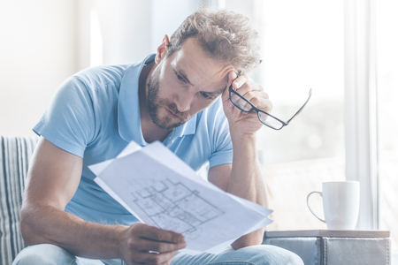Handsome White Tired Young Bearded Businessman in Casual Clothes is Examining Documents and Holding Eyeglasses in Hand while Working at Home. Work at Home. Business People Concept.の写真素材