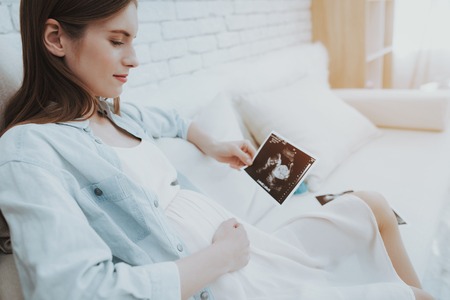 Pregnant Woman on Sofa Holds Ultrasound Scan. Happy Motherhood Concept. Healthy Pregnancy Concept. Young Mother. Expecting of New Life. Maternity Concept. Smiling Girl. Medical Photo.の写真素材