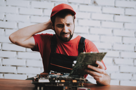 Young Man in Red Cap with Broken Motherboards in Workshop. Modern Workshop Concept. Manufacturing Instrument. Modern Tools Concept. Electronic Devices Concept. Mobile Device Hardware. Wooden Desk.の写真素材