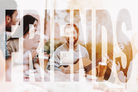 Young Guy Plays Guitar On Friends BBQ Vacation. Smiling Friends Are Having Fun. Sweater Weather. Sunny Day. Nature Relaxing. Holiday Company Meeting. Resting Together. Grill Liesure.の写真素材