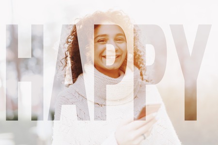 Young Female With Curly Hair. Sunny Day. Open Air Concept. White Poncho Wearing. Mulatto Girl. Mobile Phone Texting. Spring Sunlight. Natural Portrait. Sweater Weather. Pretty Cheerful Woman.の写真素材