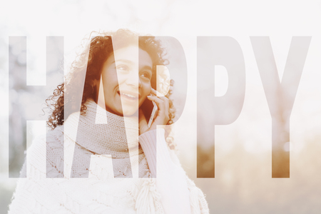 Young Female With Curly Hair. Sunny Day. Open Air Concept. White Poncho Wearing. Mulatto Girl. Mobile Phone Texting. Spring Sunlight. Natural Portrait. Sweater Weather. Pretty Cheerful Woman.の写真素材