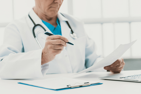 Closeup Male Doctor filling Medical Form in Office. Close up of Senior Doctor wearing White Coat and Stethoscope Sitting at Desk and Filling Examination Form in Hospital. Medicine Conceptの写真素材
