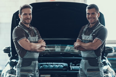 Two Mechanics Standing Near Car with Crossed Arms in Garage. Portrait of Caucasian Handsome Happy...